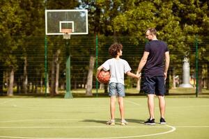 Back view full length portrait of mature man and boy standing on sports field with basket ball, holding hands photo