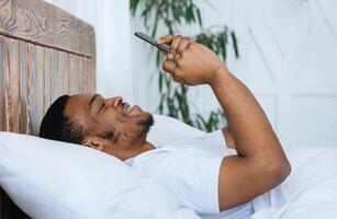Cheerful African American Guy Using Smartphone Browsing Internet On Cellphone Lying On Back In Bed At Home. Weekend Leisure, Mobile Application For Phone Concept. Side View photo