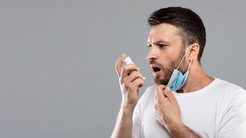 Profile portrait of man doing inhalation. Using inhaler treatment. Sick man inhaling through inhaler, hard to breath through protective face mask during pandemic. Grey studio background, copy space photo