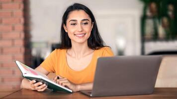 Pretty arab woman looking for job while resting at cafe, using laptop and taking notes photo