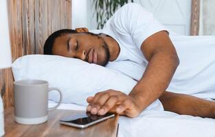 Oversleeping. Asleep African Man Sleeping Through Alarm Clock On Smartphone Waking Up In The Morning Lying In Bed In Bedroom At Home. Late For Work, Early Awakening Concept. Selective Focus photo