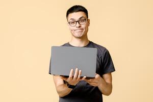 Problems at work, difficulties. Frustrated young guy student with glasses looking at camera and holds laptop, isolated on beige background, studio shot, free space photo