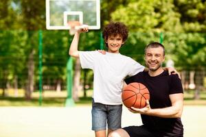 Strength Concept. Happy boy showing biceps muscles on playing basketball field, posing with his dad at camera photo
