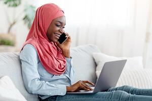 Young Black Muslim Lady In Hijab Working Remotely With Laptop And Cellphone At Home, Talking On Phone With Customer, Browsing Internet, Typing On Computer Keyboard, Sitting On Couch In Living Room photo