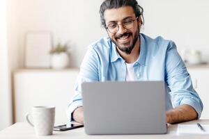 Freelance Work. Happy Millennial Arab Man Working On Computer At Home Office, Sitting At Desk With Laptop, Handsome Eastern Guy Looking At Device Screen And Smiling, Enjoying Remote Job, Copy Space photo