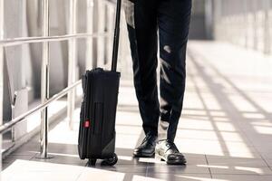 Unrecognizable businessman with luggage walking by airport, using smartphone, copy space. Cropped of man in black suit rolling suitcase and checking emails on mobile phone photo