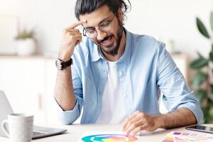 Cheerful Arab Freelance Designer Working With Color Samples At Home Office. Smiling Eastern Man Sitting At Desk With Laptop, Choosing Palette For New Design Project, Enjoying Remote Work, Free Space photo