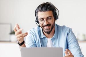 Web Conference. Positive Eastern Guy In Headset Making Call Via Laptop, Sitting At Desk In Home Office, Enjoying Online Communication, Talking And Gesturing At Device Camera, Free Space photo
