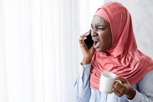 Black Muslim Woman In Headscarf Arguing On Phone And Drinking Coffee Near Window At Home, Received Bad News, Having Unpleasant Conversation, Complaining About Something, Copy Space photo