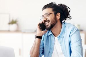 Cheerful Young Arab Guy Talking On Cellphone While Working On Laptop In Home Office. Positive Freelancer Guy Enjoying Mobile Communication And Remote Work, Sitting At Desk With Computer, Free Space photo