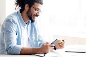 Young arab guy using smartphone and credit card at home, shopping online with cellphone and mobile app, smiling eastern man enjoying easy payments and purchases in internet, side view with copy space photo