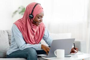 Online Education. Young Black Muslim Lady In Hijab And Headset Studying With Laptop Computer At Home, Taking Notes While Watching Webinar, Enjoying Distance Learning, Sitting On Couch, Copy Space photo