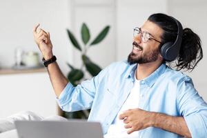 Be Yourself. Joyful weatern freelancer guy listening music in headphones and playing virtual guitar, relaxing at desk in home office, young indian man having fun and enjoying working remotely photo