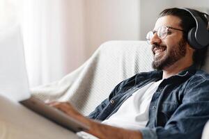 Indian Freelancer Man Working On Laptop Online At Home And Listening Music With Wireless Headphones, Making Research For New Project, Sitting On Couch With Computer, Enjoying Remote Job, Copy Space photo