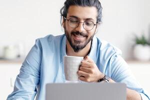 Young Smiling Arab Freelancer Guy Using Laptop And Drinking Coffee At Home, Relaxing With Hot Drink While Working Remotely Online On Computer, Enjoying Distance Job Opportunities, Free Space photo