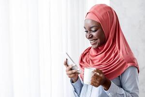 Relaxing Morning. Smiling Black Muslim Woman In Hijab Drinking Coffee And Using Smartphone At Home, Standing Near Window, Enjoying Hot Drink And Mobile Communication, Reading Messages, Copy Space photo