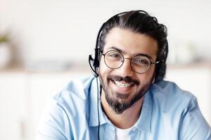 Call Center Operator. Closeup portrait of positive indian man customer service representative in headset, handsome western guy hotline manager looking at camera and smiling, selective focus photo