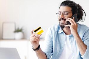 Easy Payments. Happy Arab Freelancer Guy Holding Credit Card And Talking On Cellphone While Sitting At Desk In Home Office, Making Mobile Payment, Speaking With Customer Service Manager, Free Space photo