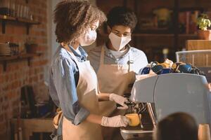 Barista work during social distancing and covid-19 pandemic. Millennial african american lady and guy in aprons and protective masks use coffee machine behind bar in cafe interior, empty space photo