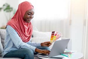 Black Muslim Woman Designer Using Laptop And Holding Color Swatch At Home, Working Remotely In Internet, Studying Online, Enjoying Distance Learning, Sitting On Couch In Living Room, Copy Space photo