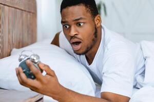 Oversleeping Concept. Shocked Black Guy Looking At Broken Alarm Clock Waking Up Late For Work In The Morning, Lying In Bed At Home. Awakening Problem and Tardiness, Bad Morning photo