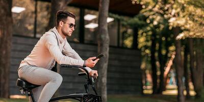 Tries to find his best road during stop with gps online map. Concentrated handsome millennial man in sunglasses looks at smartphone, sits on modern bicycle in park, outdoor, side view, free space photo