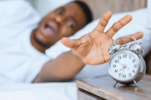 Early Wake-Up. Stressed African Man Shouting Turning Off Alarm-Clock Waking Up Too Early Lying In Bed At Home. Shallow Depth photo