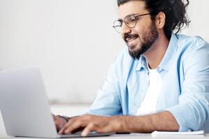 Busy Smiling Indian IT Manager Guy Working Online On Laptop At Workplace In Office, Typing On Keyboard, Cheerful Western Male Freelancer In Eyeglasses Using Computer, Browsing Internet, Free Space photo