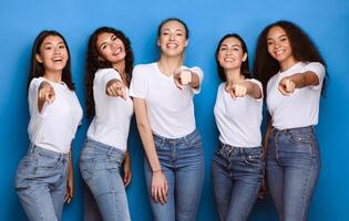 Five Multiethnic Women Pointing Finger At Camera Choosing You Posing Standing Over Blue Studio Background. You're Next Concept. Advertisement Banner With Cheerful Diverse Ladies photo