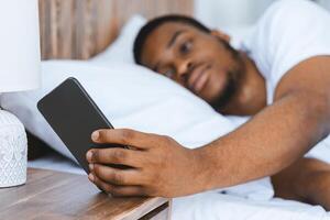 Black Man Turning Off Alarm Clock On Smartphone Waking Up Early In The Morning Lying In Bed At Home. Lack Of Sleep, Rest And Recreation Concept. Selective Focus, Shallow Depth photo