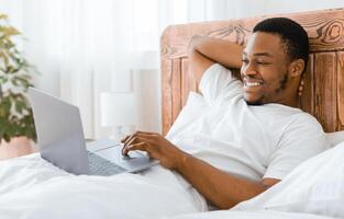 Cheerful African American Man Using Laptop Watching Movie Online And Browsing Internet Lying In Bed At Home On Weekend Morning. Computer User Searching Entertaining Web Content Concept photo