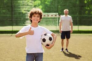 Handsome curly boy holding soccer ball on the field, showing thumb up, man standing in the blurred background photo