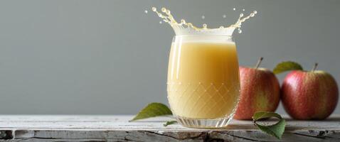 A glass of apple juice with a splash surrounded by two apples on a wooden surface and gray background photo