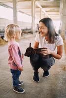 Smiling mother, squatting down, shows her little daughter a black goatling in her arms photo