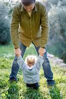 Dad stands over a little girl sitting on the lawn and holds her hands photo