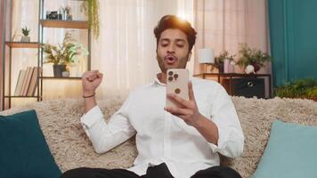 Indian joyful man on sofa happily texting on smartphone, celebrating lottery win, joyful expression video