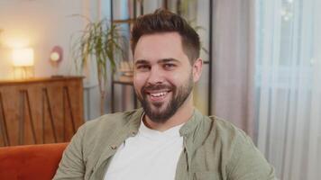 Portrait of happy young Caucasian man with brown hair looking at camera sitting on sofa at home video