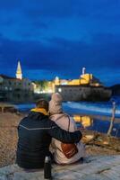 Man and woman sit embracing on the seashore and look at the illuminated old houses. Back view photo