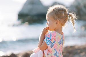 Little girl is standing on the beach with a soft toy and chewing on her finger. Side view photo