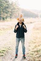 Smiling dad with a little son on his shoulders stands in a sunny forest photo