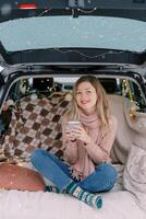 Smiling girl with a cup of coffee sitting cross-legged in the trunk of a car on a blanket photo