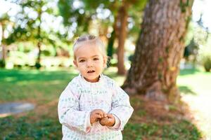 Little smiling girl stands near a tree in a sunny forest with a nut in her hands photo
