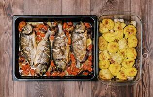 Baked sea bream with vegetables on a baking sheet stands with baked potatoes in a dish on a wooden table. Top view photo