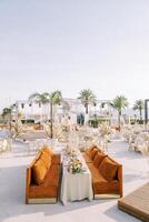 Sofas with cushions stand along a long covered table surrounded by round tables on the seashore photo