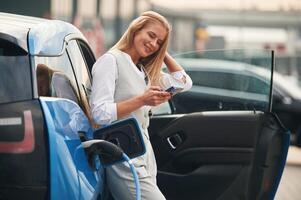 Smartphone in hands. .Woman ,on the,electric cars charge station at daytime photo