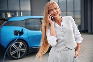 Using, smartphone,. Woman on the electric cars charge station at daytime photo