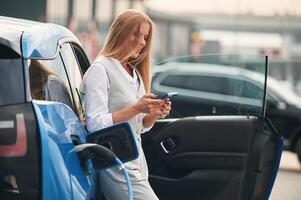 Smartphone in hands.. Woman on the electric cars charge station at daytime photo