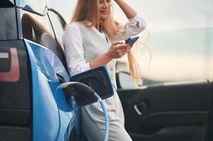 Smartphone in ,hands. Woman on the electric cars charge station at daytime photo