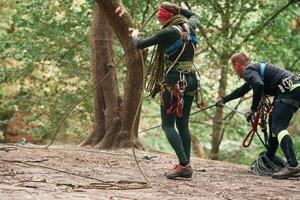Standing, preparing the ropes. Man and woman doing climbing in forest with use of safety equipment photo