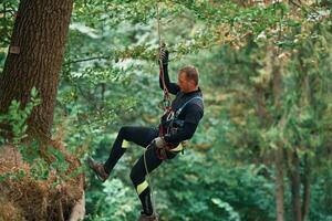 Hanging on a, rope. Man is, doing climbing in the forest by use of safety equipment photo